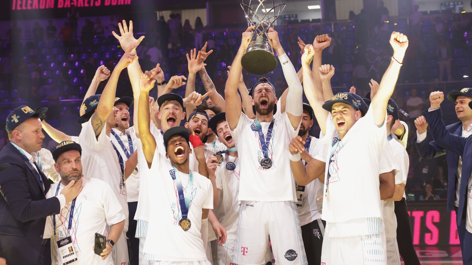 Männer mit Medaillen um den Hals jubeln. Eine Person hält beidhändig einen Pokal in die Luft.