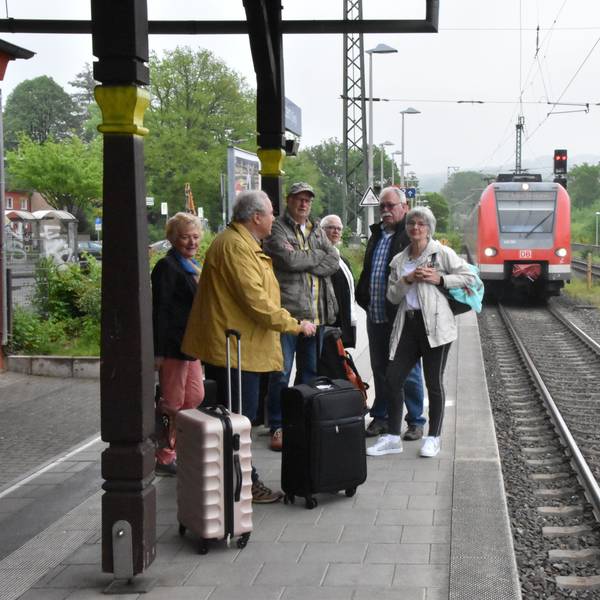 Auf einem Bahnsteig steht eine Gruppe Menschen mit Koffern, ein Zug fährt ein.