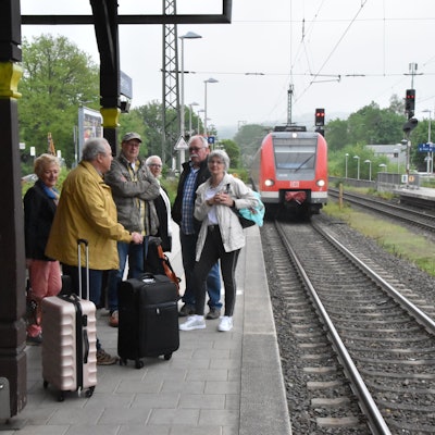 Auf einem Bahnsteig steht eine Gruppe Menschen mit Koffern, ein Zug fährt ein.