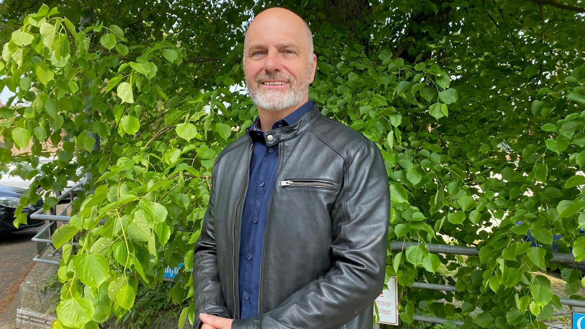 Marcus Venghaus trägt einen Ledermantel und steht vor einem grünen Baum.