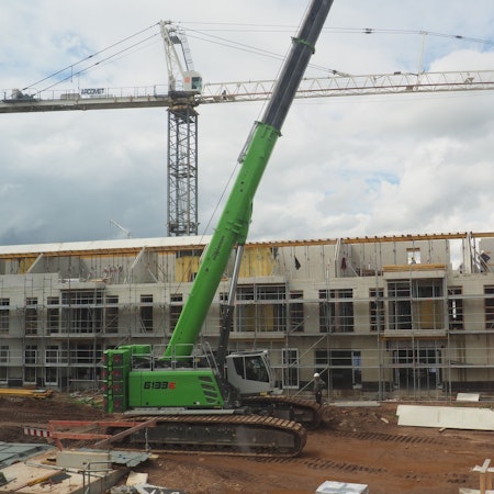 Ein grünes Baustellenfahrzeug steht vor eine Reihe von Häusern im Rohbau. Dahinter ist ein Kran zu sehen.