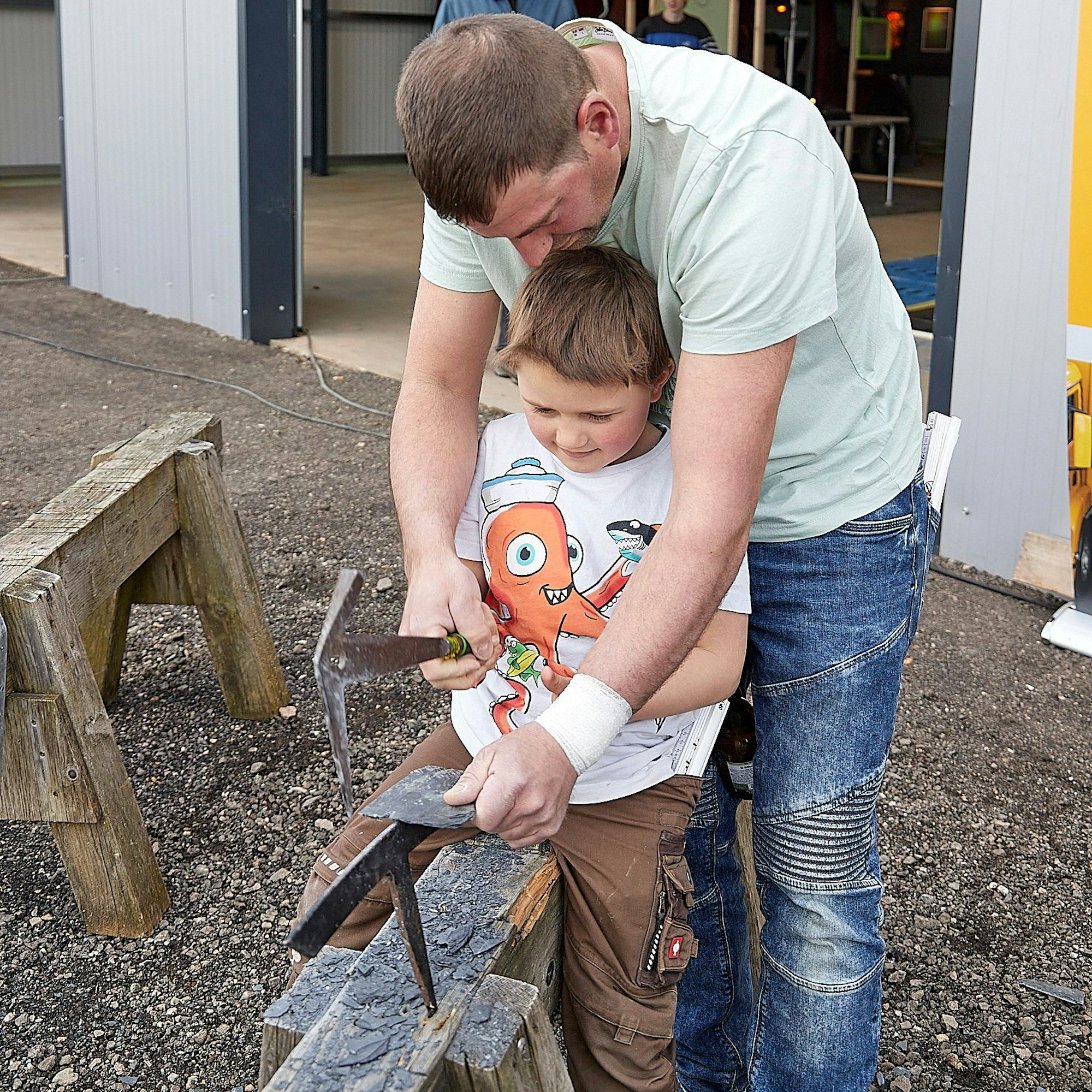 Der Vater zeigt dem Jungen, wie mit einem speziellen Hammer auf einem Holzbock der Schiefer in Form gebracht wird.