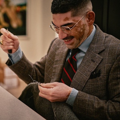 Ein Mann im Anzug sitzt vor einer Nähmaschine. Er hat Nadel und Faden in der einen und ein Stück Stoff in der anderen Hand.