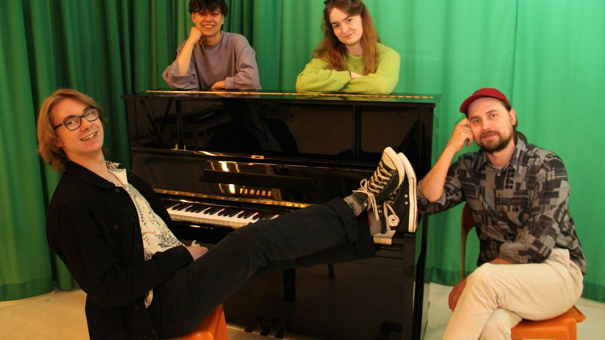 Um ein Klavier herum sitzen und stehen drei junge Erwachsene und ein älterer Mann.