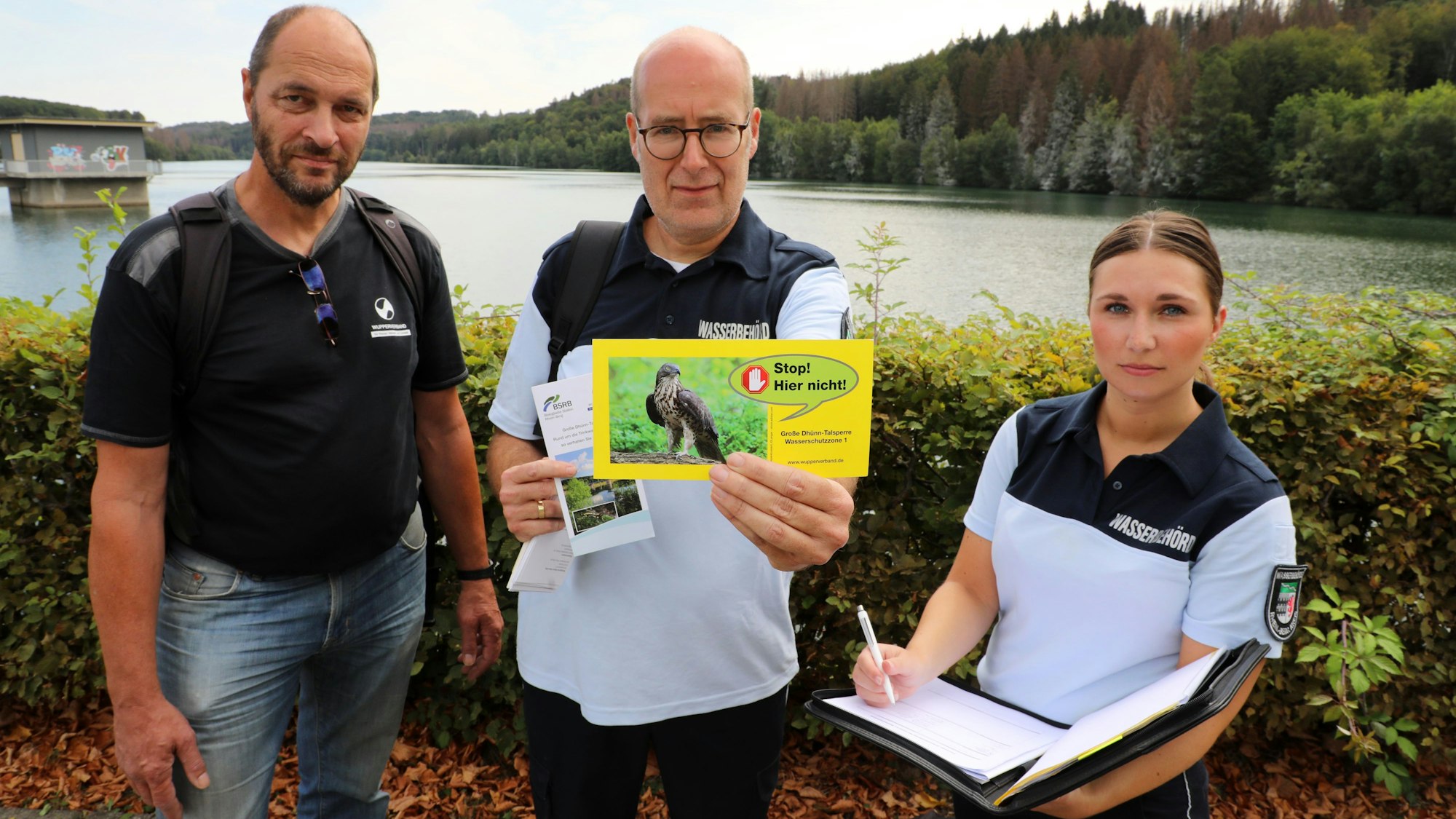Zwei Männer und eine Frau stehen in Uniform vor einer Talsperre und halten eine gelbe Karte noch.