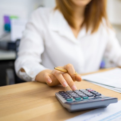 Unscharf zu sehen ist eine junge Frau am Schreibtisch, im Vordergrund ein Taschenrechner, Unterlagen und Formulare.