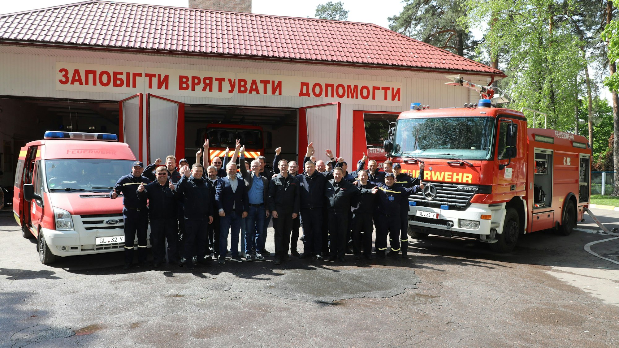 Eine Gruppe von Feuerwehrleuten und Zivilisten steh zwischen zwei Feuerwehrfahrzeugen und jubelt. Im Hintergrund steht eine Feuerwache der ukrainischen Stadt Butscha.