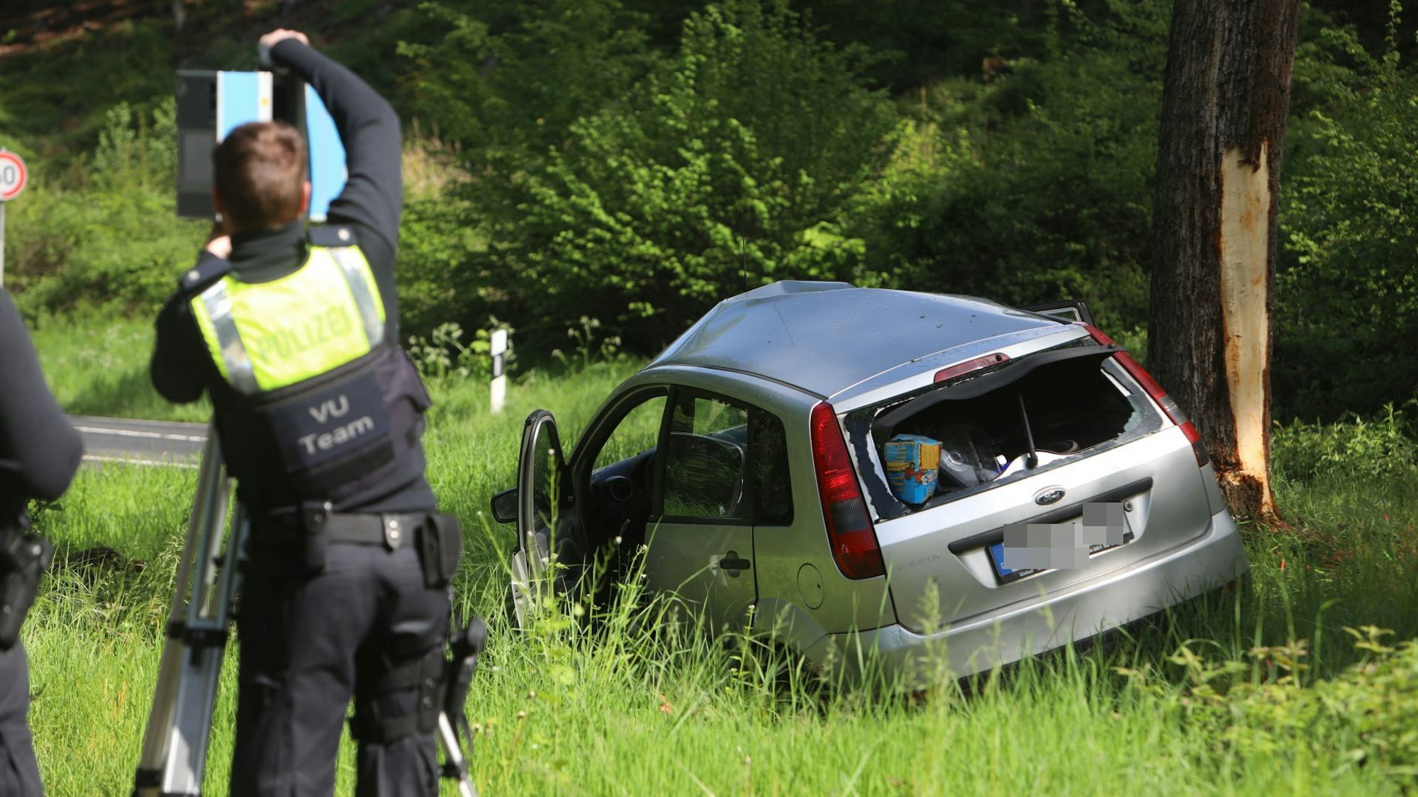 Zwei Polizisten bauen ein Stativ auf, vor ihnen im hohen Gras liegt ein verbeulter Pkw neben einem Baum, dessen Rinde abgeschält ist.