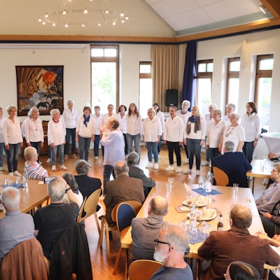 Zu sehen ist ein Frauenchor, der vor sitzenden Gästen singt.