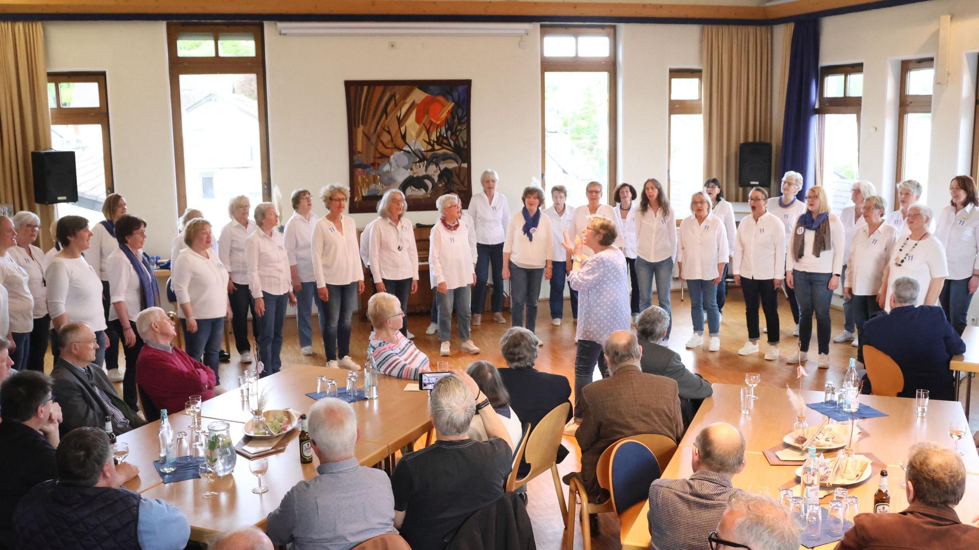 Zu sehen ist ein Frauenchor, der vor sitzenden Gästen singt.