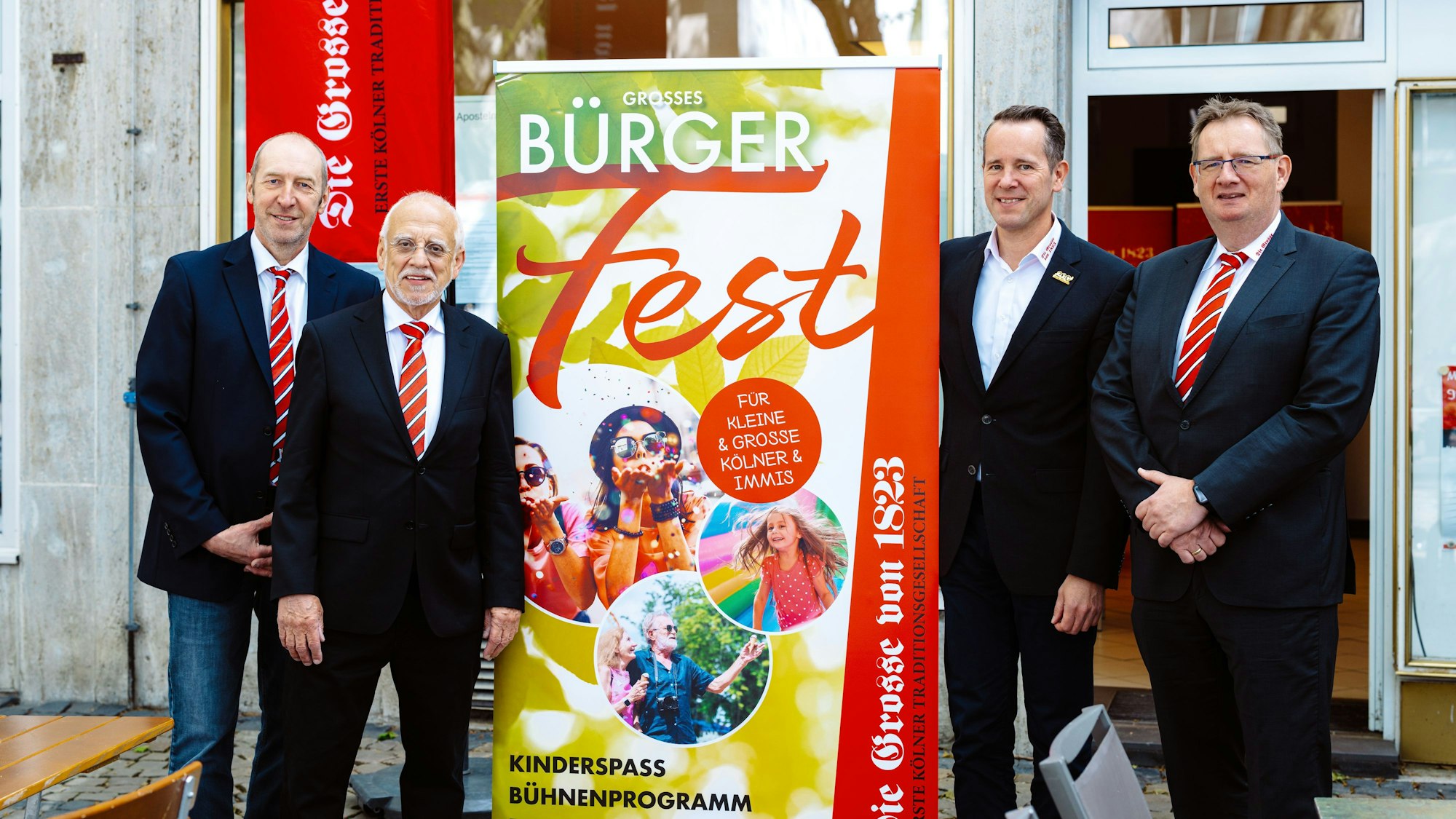 Vier Männer im Anzug stehen neben einem Plakat, das für das Bürgerfest wirbt.