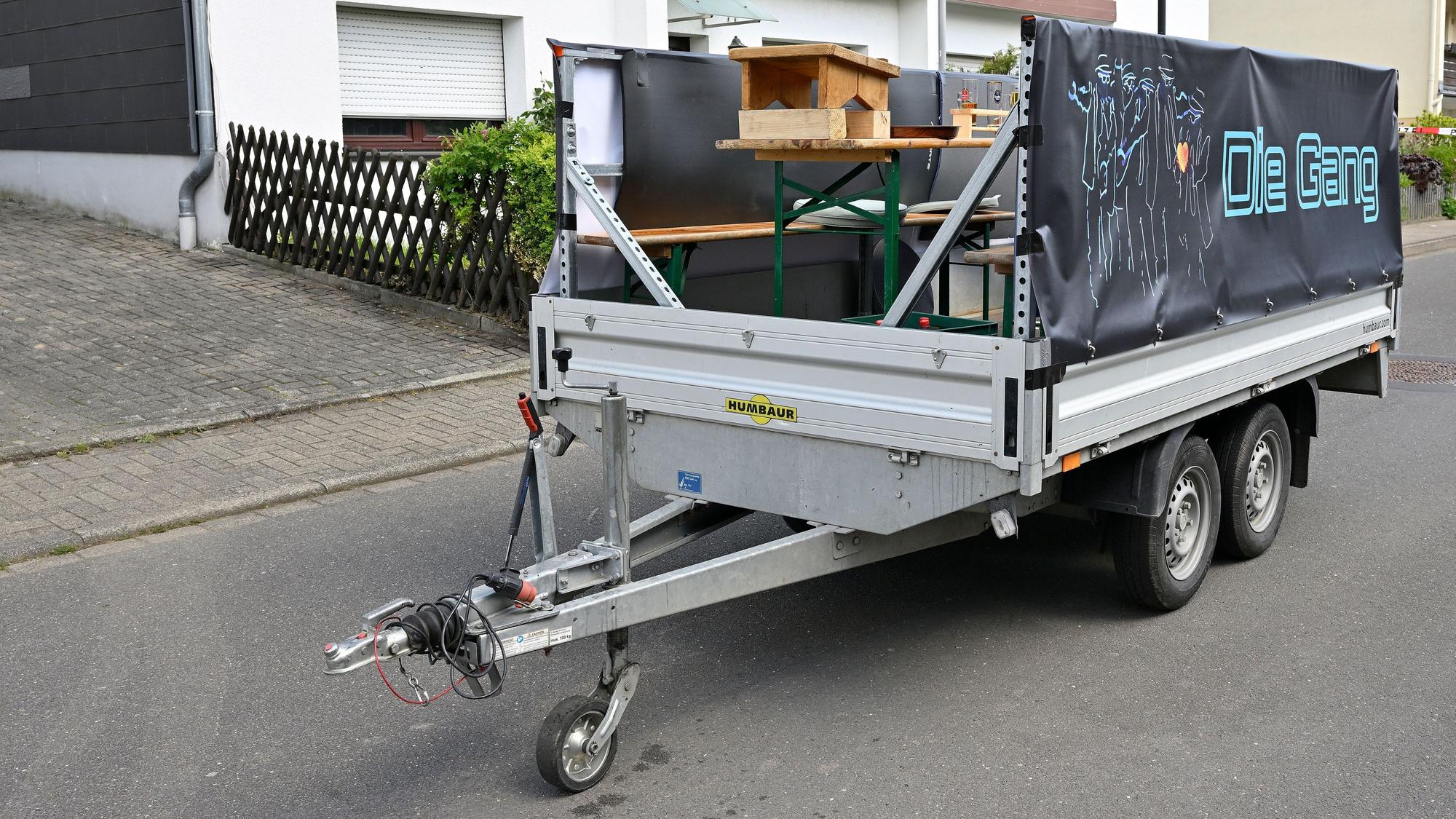 Ein Anhänger steht auf einer Straße, auf der Ladefläche befinden sich Bänke, ein Tisch und ein Podest für ein Bierfässchen..