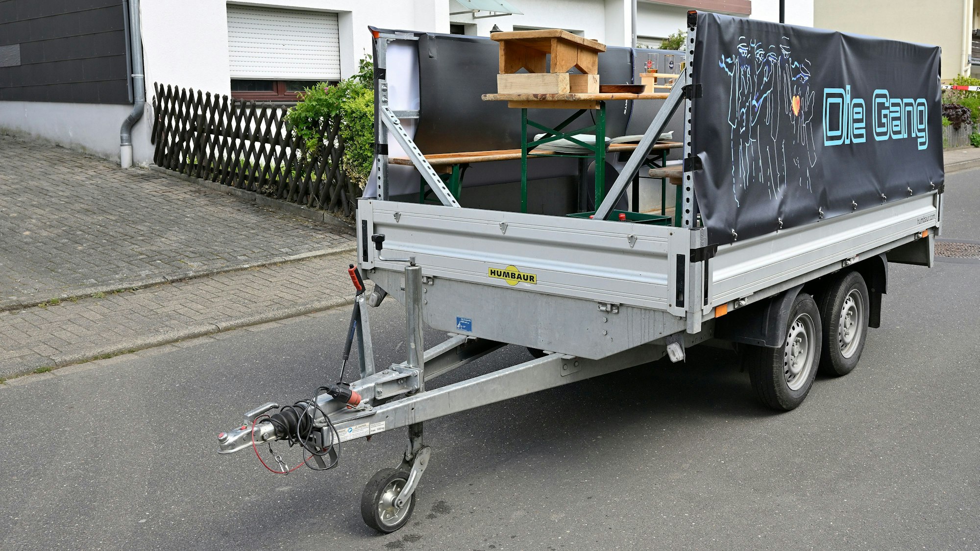 Ein Anhänger steht auf einer Straße, auf der Ladefläche befinden sich Bänke, ein Tisch und ein Podest für ein Bierfässchen..