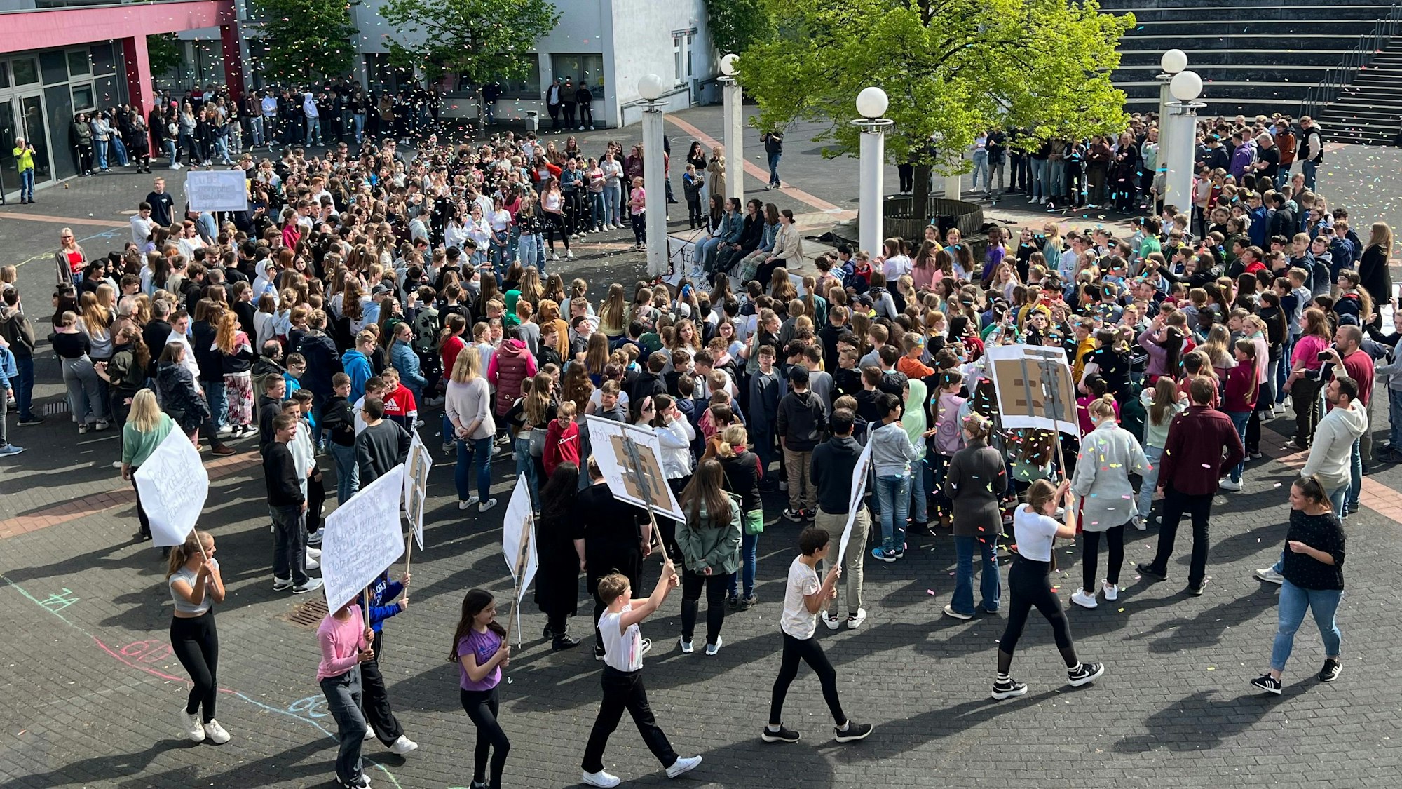 Eine Traube an Menschen versammelt sich auf einem Schulhof. Einige halten Plakate in die Höhe.