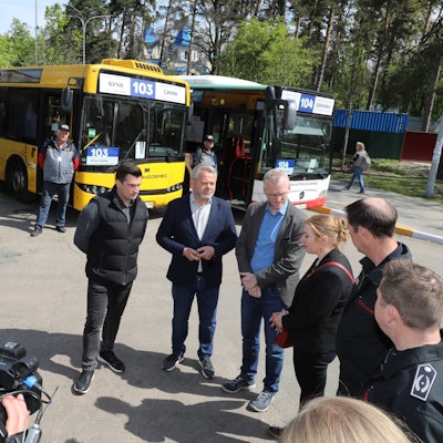 Menschen stehen im Halbkreis vor einer Reihe von Bussen. Ein Kameramann hält das Geschehen fest.