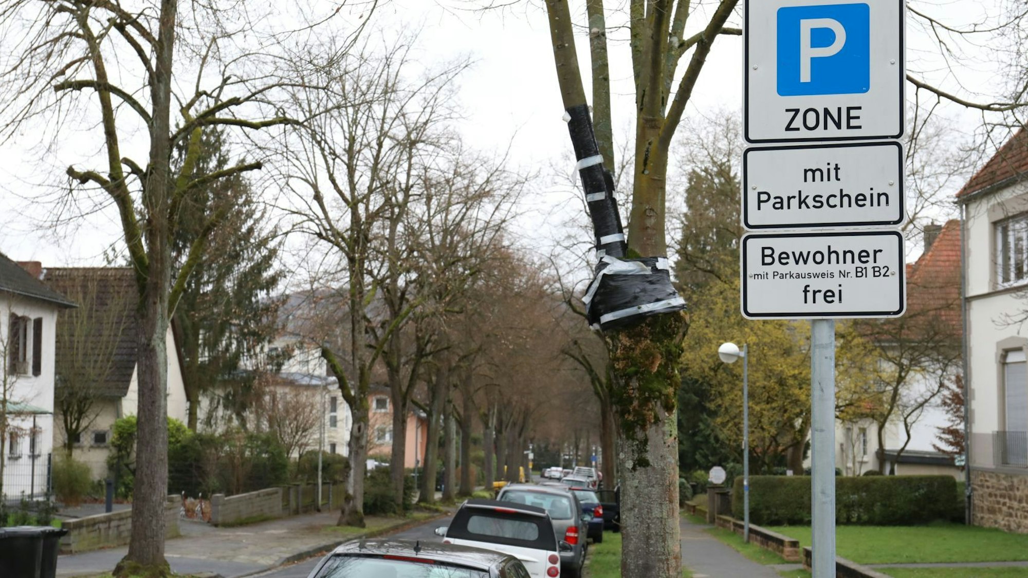 Ein Schild weist die Straße als Parkzone mit Parkschein aus, Bewohner mit Ausweis können dagegen frei parken. In der Straße stehen rechts am Fahrbahnrand parkende Autos.