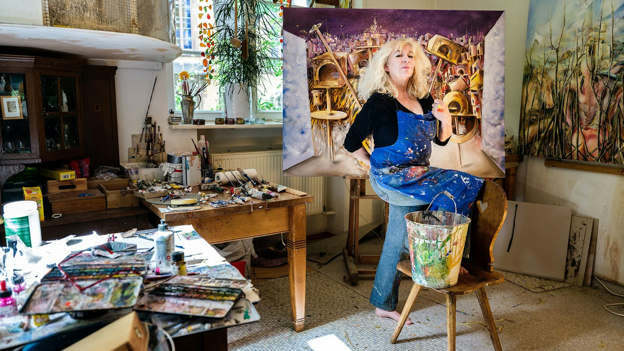 Eine Künstlerin posiert in einem Atellier vor einem bunten Bild auf einer Leinwand.