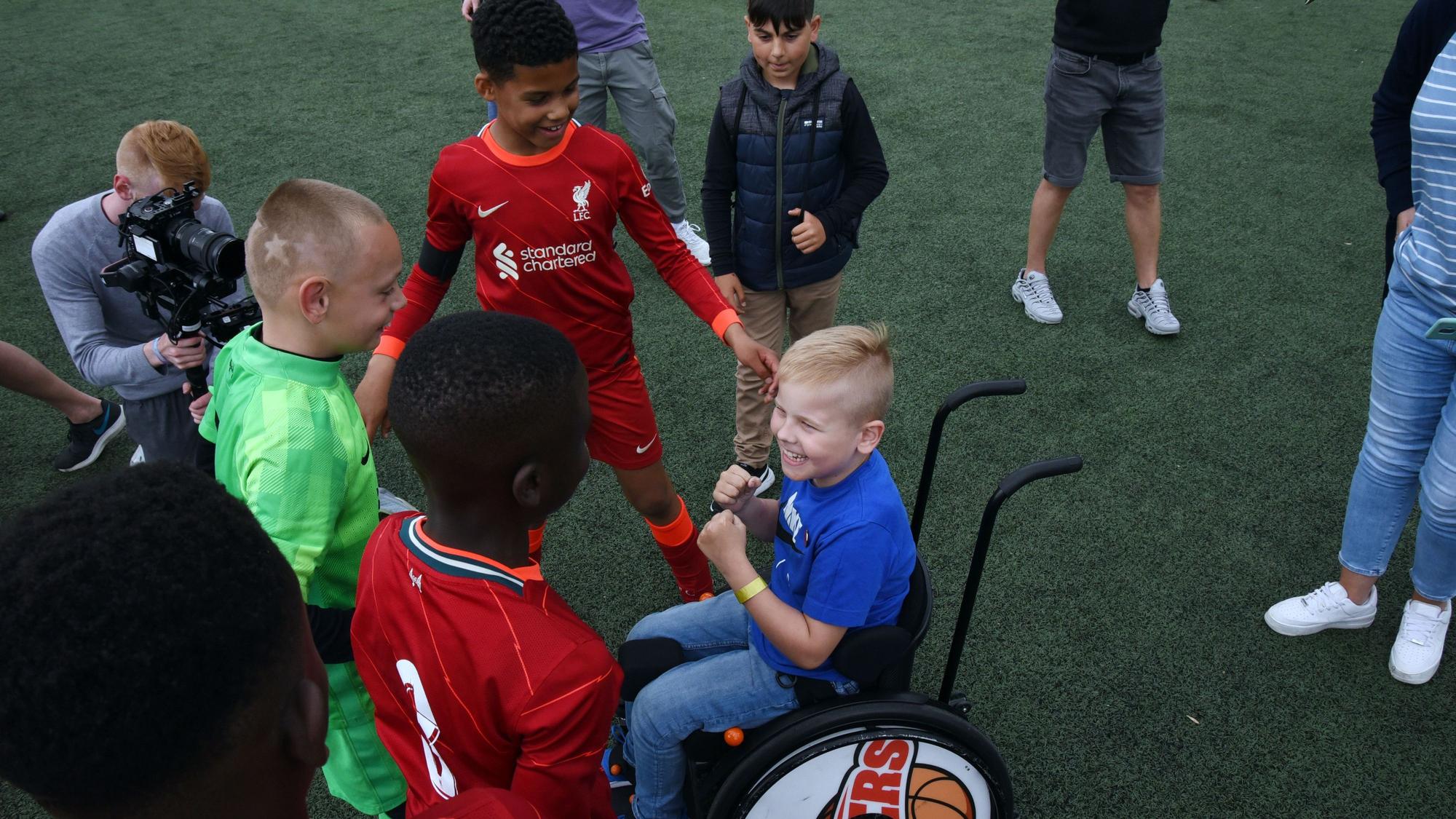 Mehrere haben sich auf einem Fußballplatzversammelt und lachen. Eines der Kinder sitzt in einem Rollstuhl.