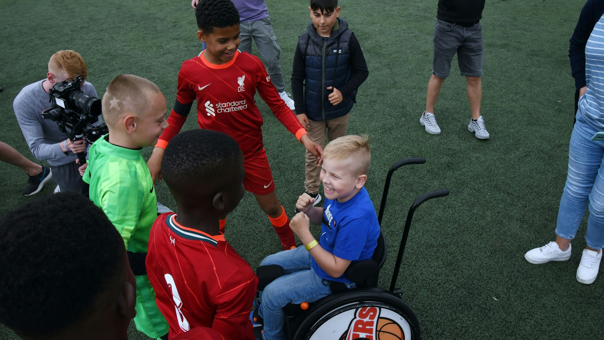 Mehrere haben sich auf einem Fußballplatzversammelt und lachen. Eines der Kinder sitzt in einem Rollstuhl.
