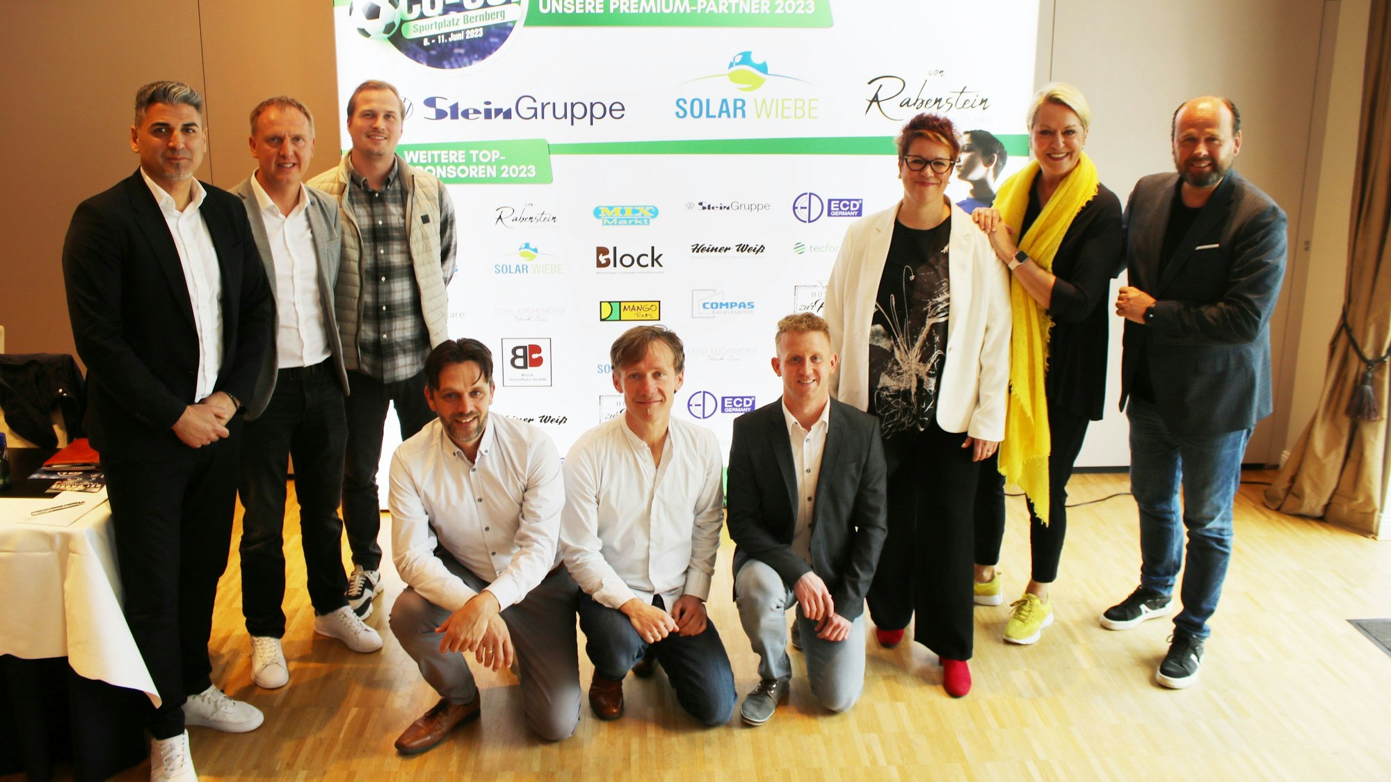 Mehrere Männer und Frauen stehen vor einer Sponsoren-Tafel zusammen.