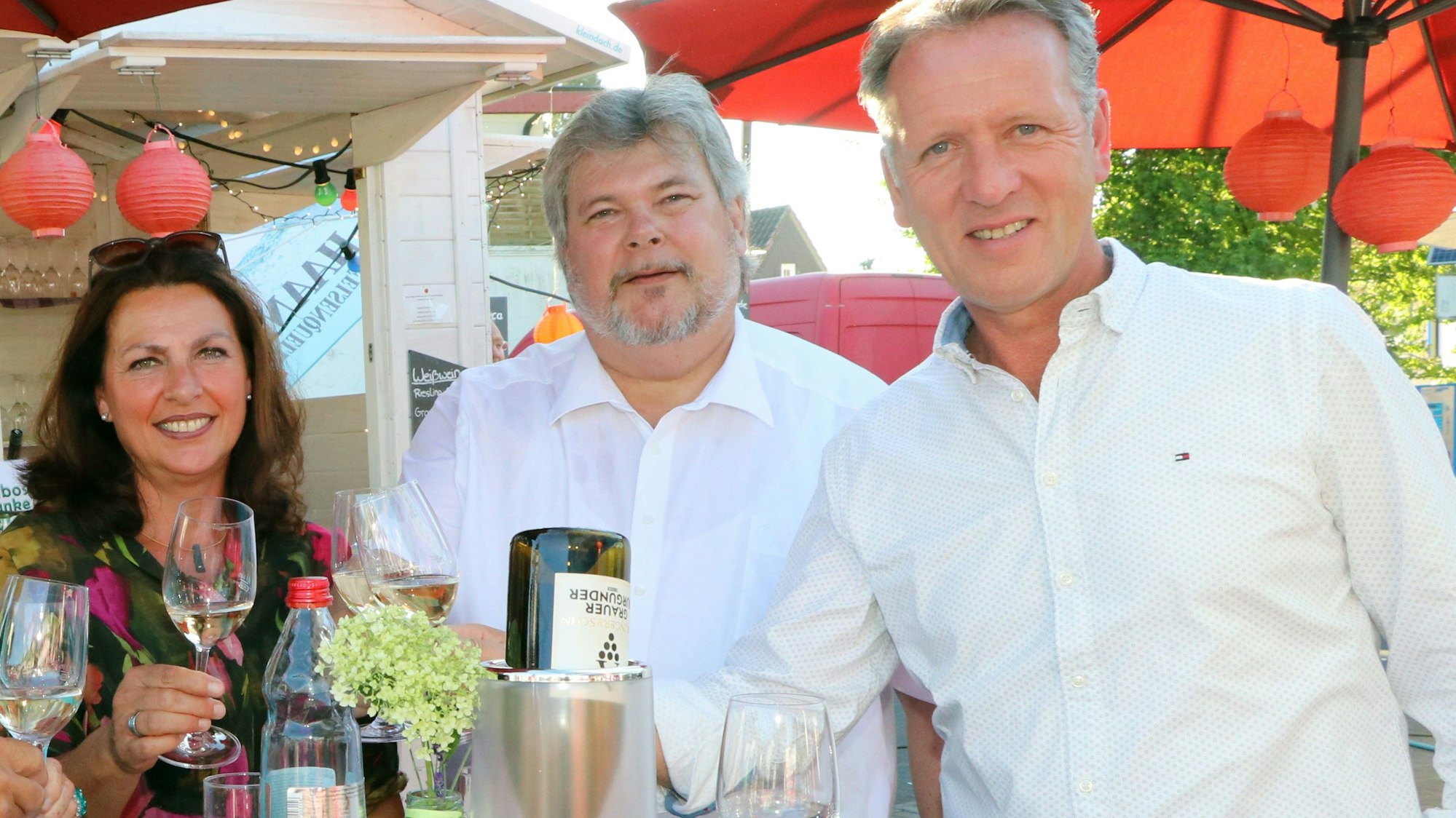 Zwei Männer und eine Frau stehen auf einem Fest an einem Stehtisch und trinken Wein.