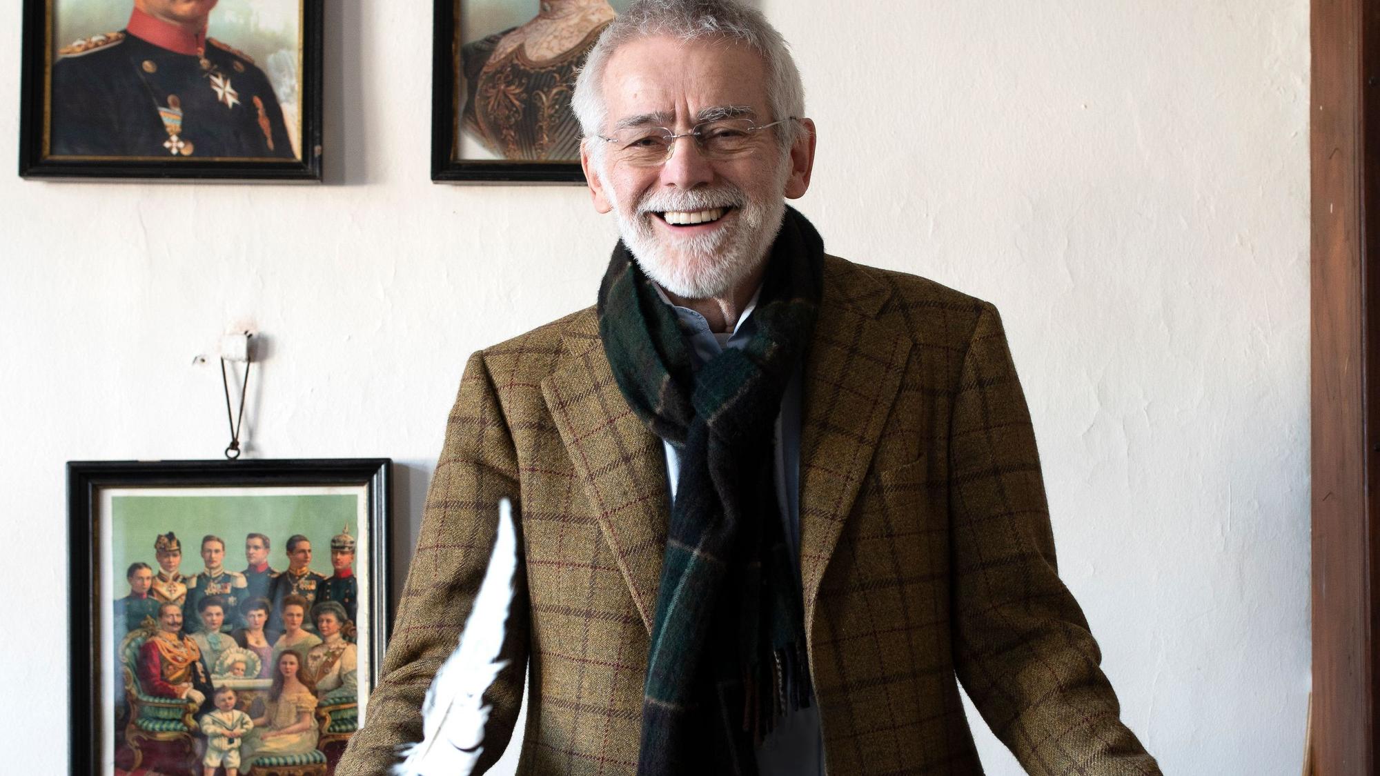 Dr. Peter Joerißen, Leiter Schulmuseum Bergisch Gladbach, im alten Klassenzimmer der ehemaligen Volksschule. Er steht vor einigen Gemälden, die an einer weißen Wand hängen, und lächelt an der Kamera vorbei.