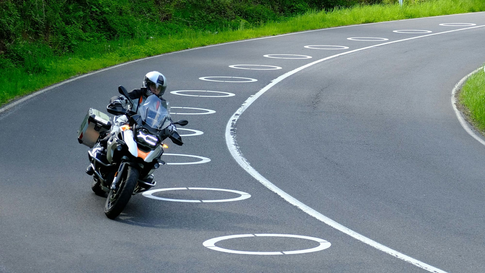 Ein Motorradfahrer umfährt mit einem Bike in einer Linkskurve auf der Fahrbahn angebrachte, ellipsenförmige Fahrbahnmarkierungen.