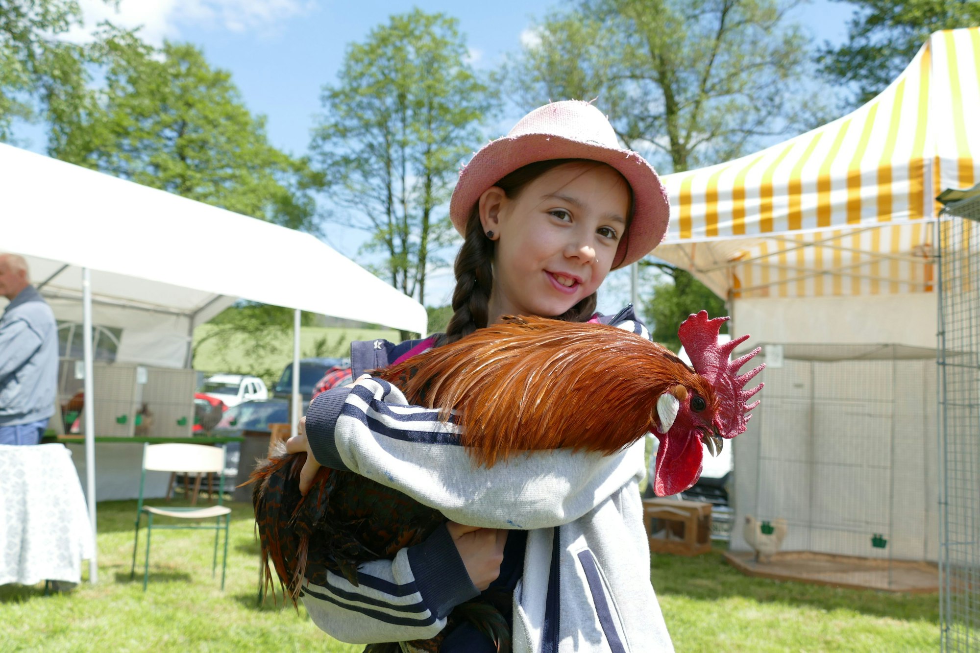 Ein Mädchen mit Hut hält fröhlich einen Hahn - im Hintergrund stehen Käfige und Zelte.