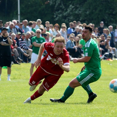 Mechernichs Jonas Hohn sprintet an Sötenichs Marvin Neumann vorbei. Im Hintergrund sieht man neben den Sötenicher Verantwortlichen viele Zuschauer.
