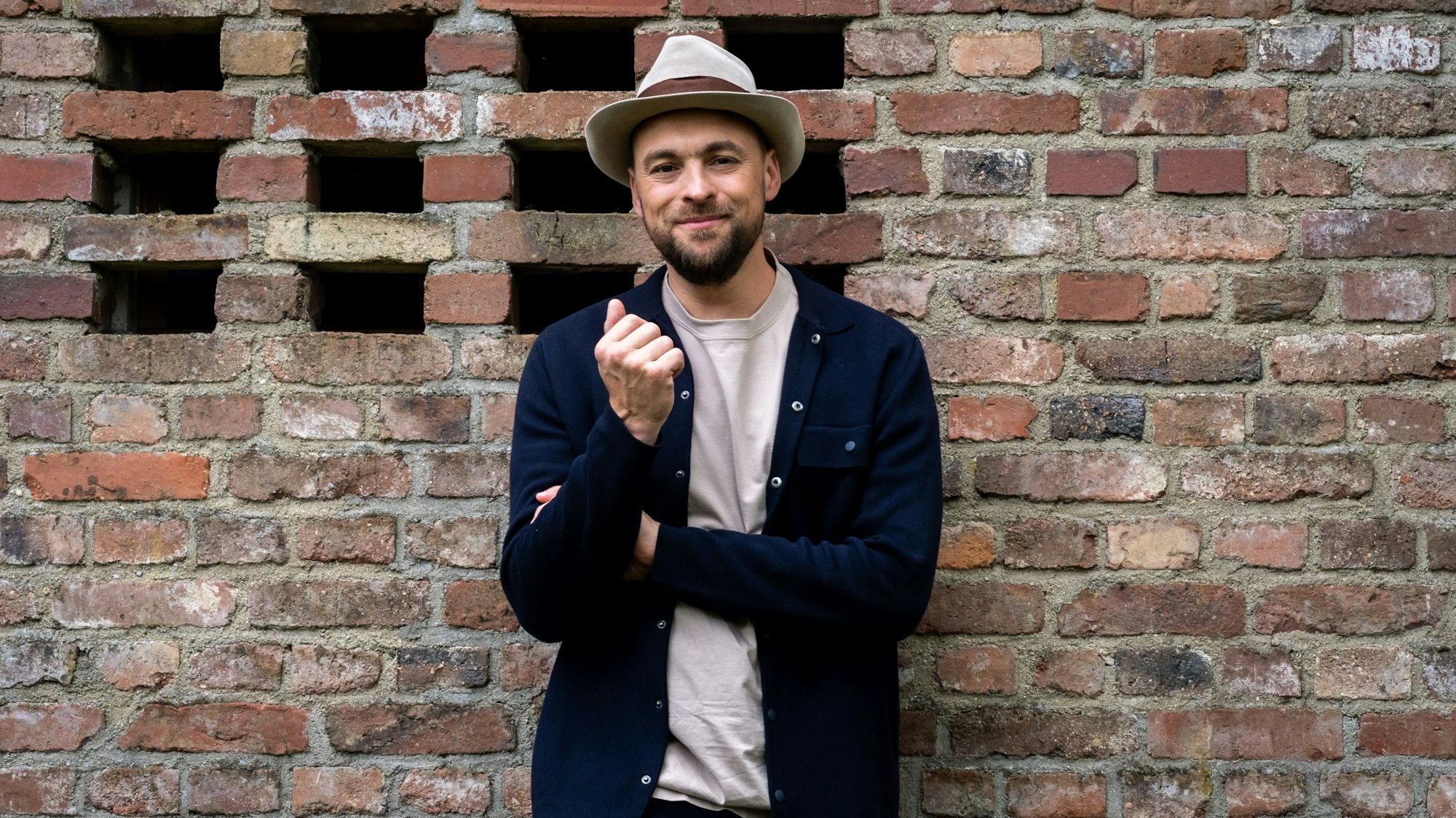 Der Sänger Max Mutzke steht lächelnd mit einem Hut auf dem Kopf vor einer Mauer.