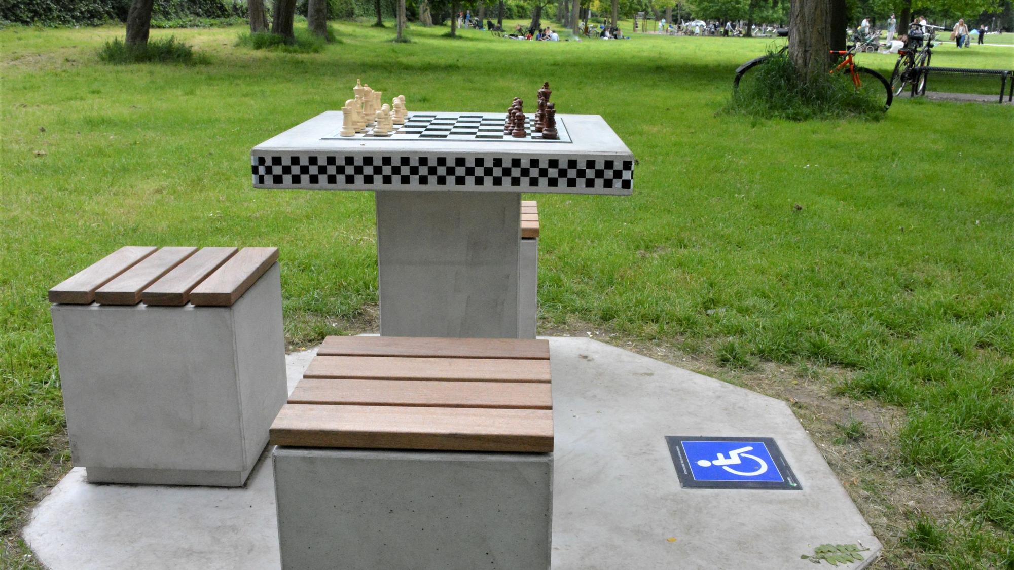 Ein Schachtisch aus Beton steht im Grüngürtel, eine Aufstellfläche für einen Rollstuhl wird mit einem Signet angezeigt.