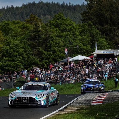 Im Bild sind zwei Rennwagen, die hintereinander fahren, dahinter sind Tribünen mit Zuschauern zu sehen.