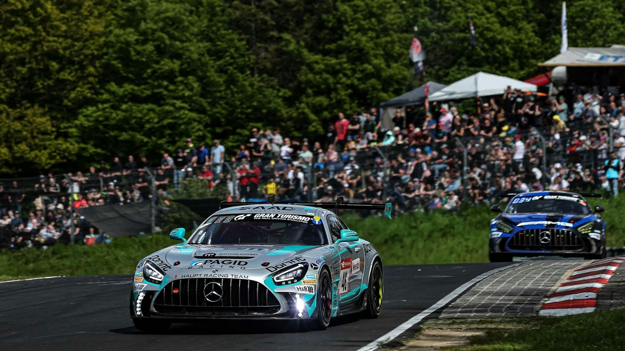 Im Bild sind zwei Rennwagen, die hintereinander fahren, dahinter sind Tribünen mit Zuschauern zu sehen.