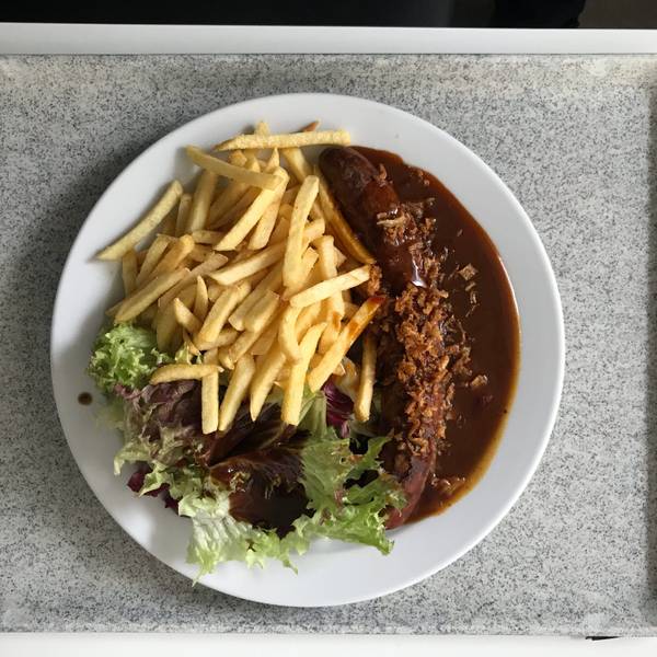 Ein Teller mit Currywurst, Pommes frites und einer kleinen Salatbeilage steht auf einem Tablet in einer Kantine.