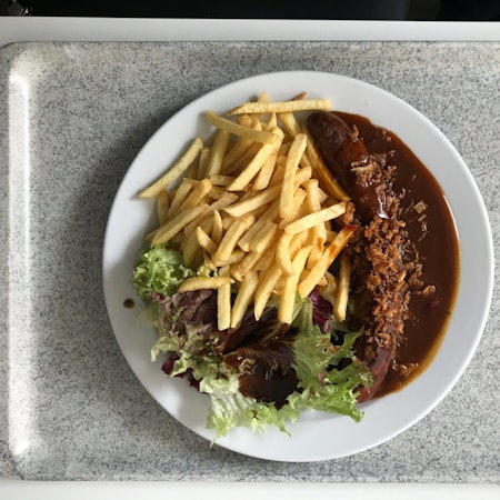 Ein Teller mit Currywurst, Pommes frites und einer kleinen Salatbeilage steht auf einem Tablet in einer Kantine.