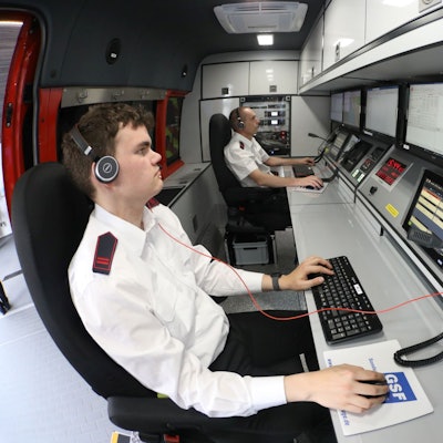Zwei Männer sitzen vor Computermonitoren im Einsatzleitwagen der Feuerwehr und bedienen Maus und Tastatur. Beide tragen Headsets.