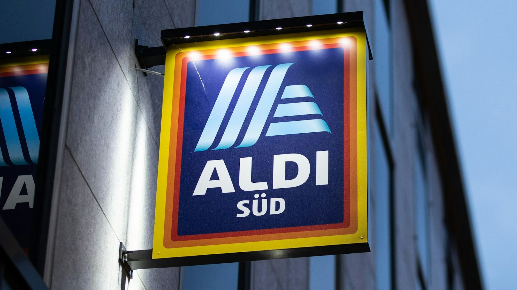 Das Logo einer Aldi Süd Filiale, fotografiert kurz vor dem Sonnenuntergang. Das Schild wird beleuchtet.