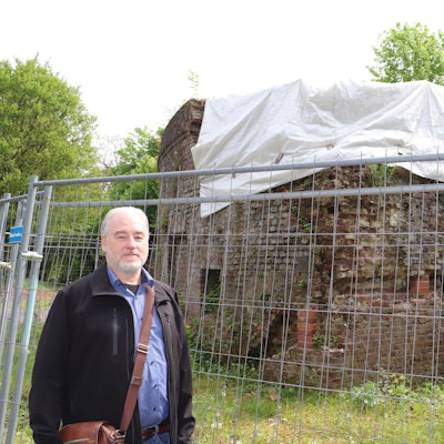 Ein Mann steht vor einem alten Gemäuer, das durch einen Bauzaun abgesperrt und das zur Hälfte mit einer weißen Plane abgedeckt ist.