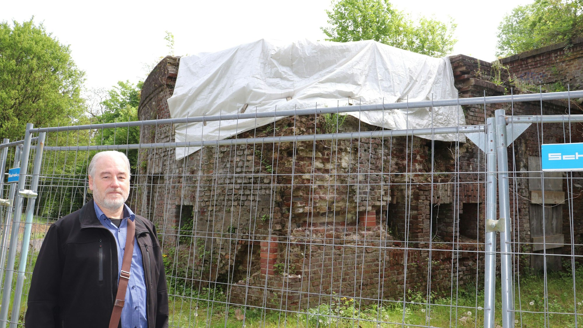Ein Mann steht vor einem alten Gemäuer, das durch einen Bauzaun abgesperrt und das zur Hälfte mit einer weißen Plane abgedeckt ist.