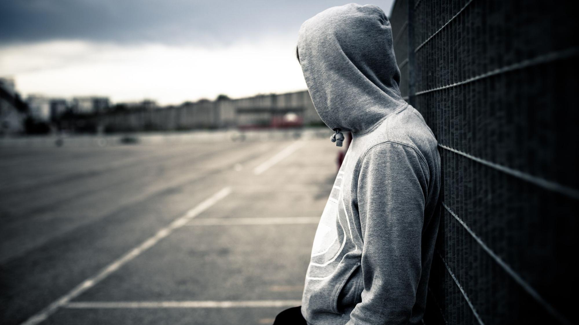 Ein junger Mann mit Kapuzenpulli lehnt alleine an einem Zaun.