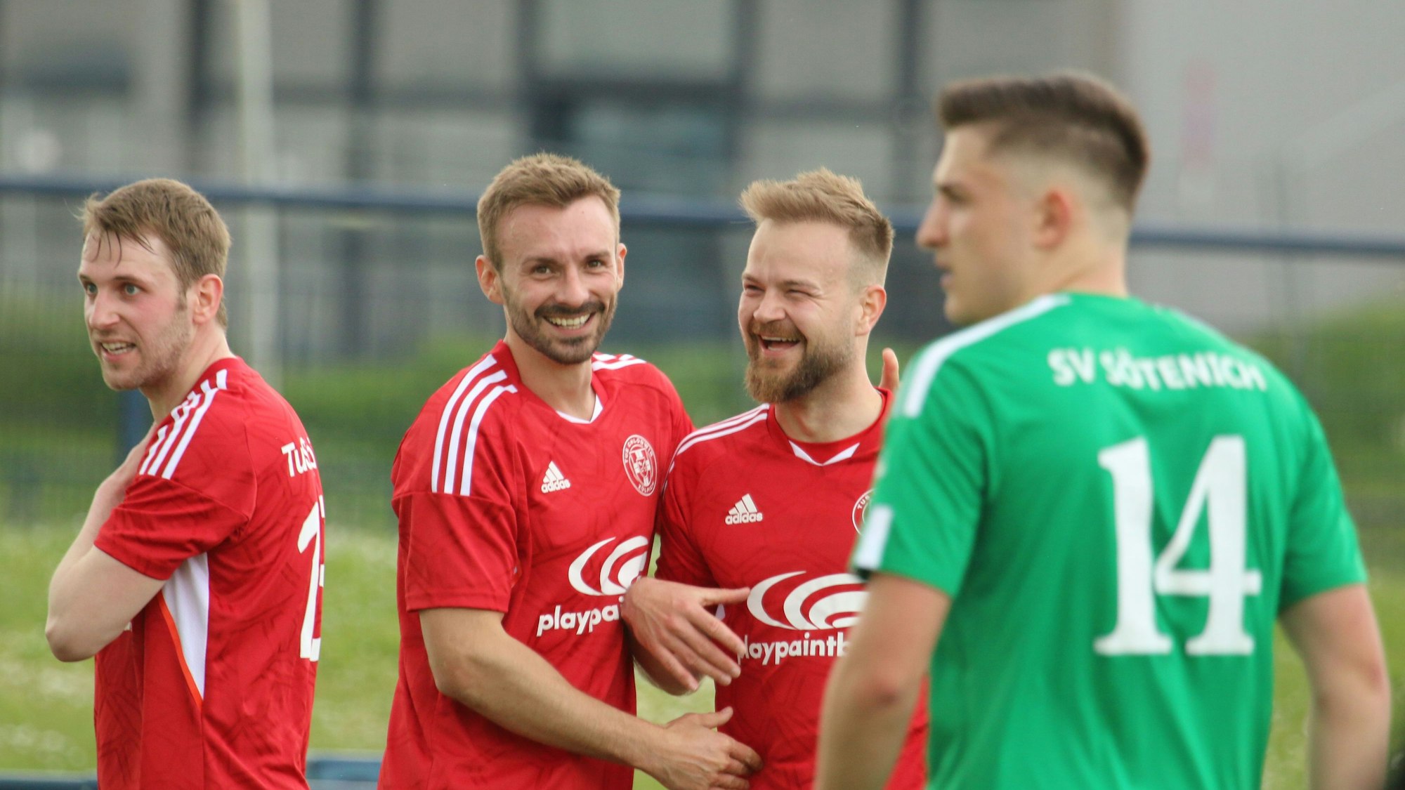 Vier Fußballspieler sind zu sehen, drei davon tragen rote Trikots und sind gut gelaunt, einer trägt grün und schaut in die Ferne.
