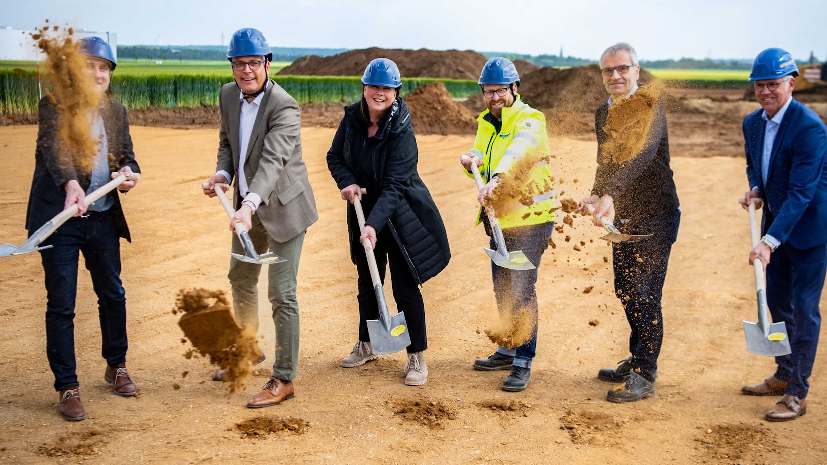 Sechs Personen stehen auf einem sandigen Untergrund und schaufeln zu einem symbolischen ersten Spatenstich ein wenig Sand.