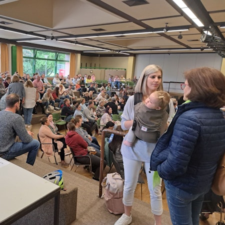 Eine Frau mit einem Kind in der Trage steht in einem Saal mit vielen Menschen.