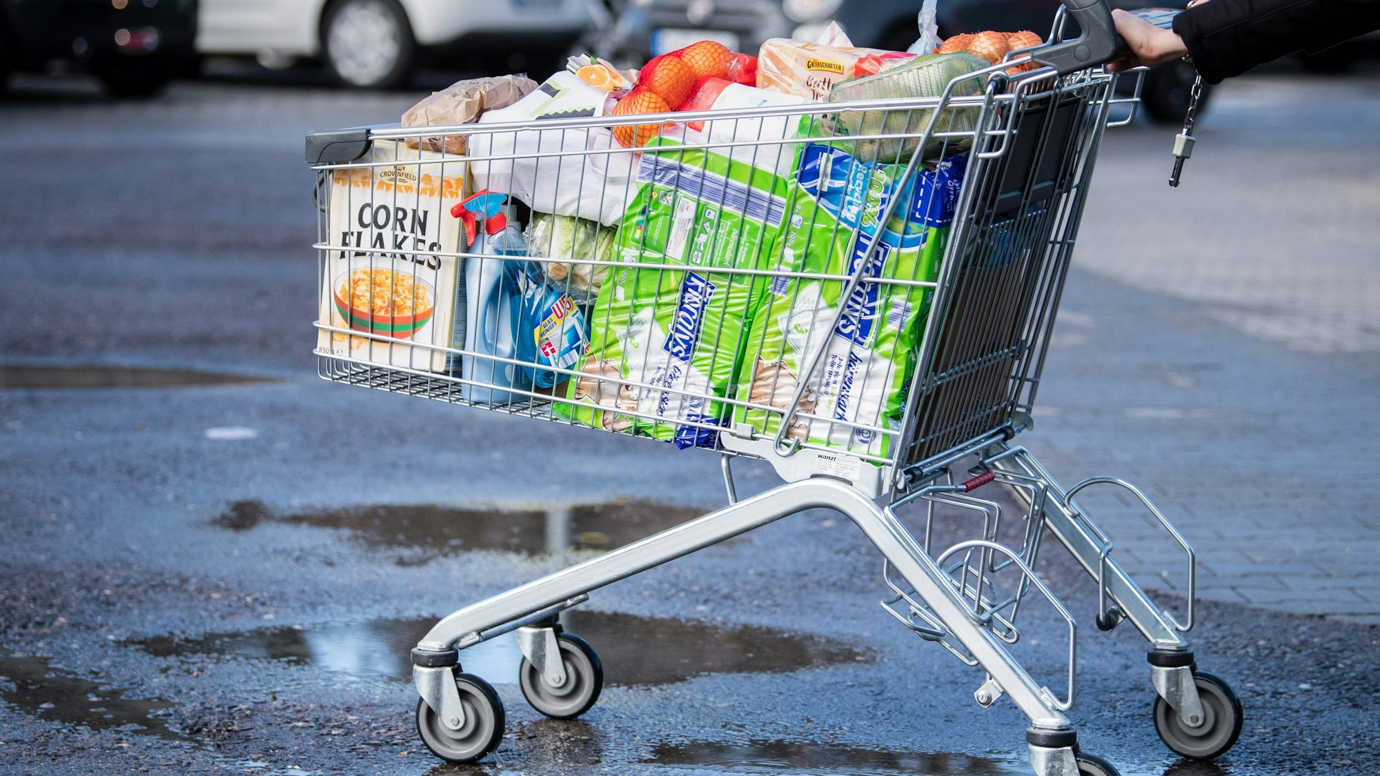 Ein Kunde schiebt einen bis zum Rand gefüllten Einkaufswagen über den Parkplatz eines Supermarktes.