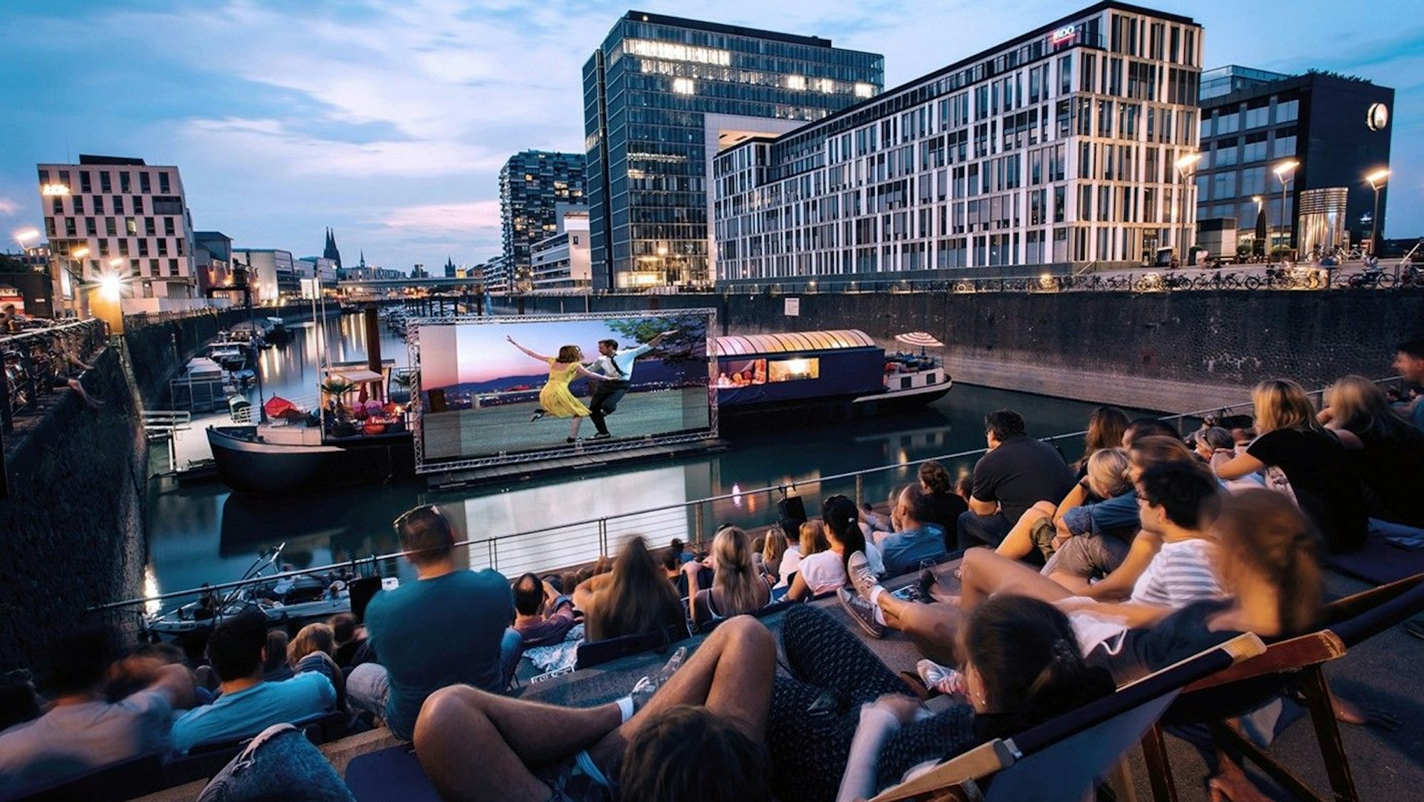 Gäste sitzen vor einer bespielten Leinwand im Sommerkino im Rheinauhafen.