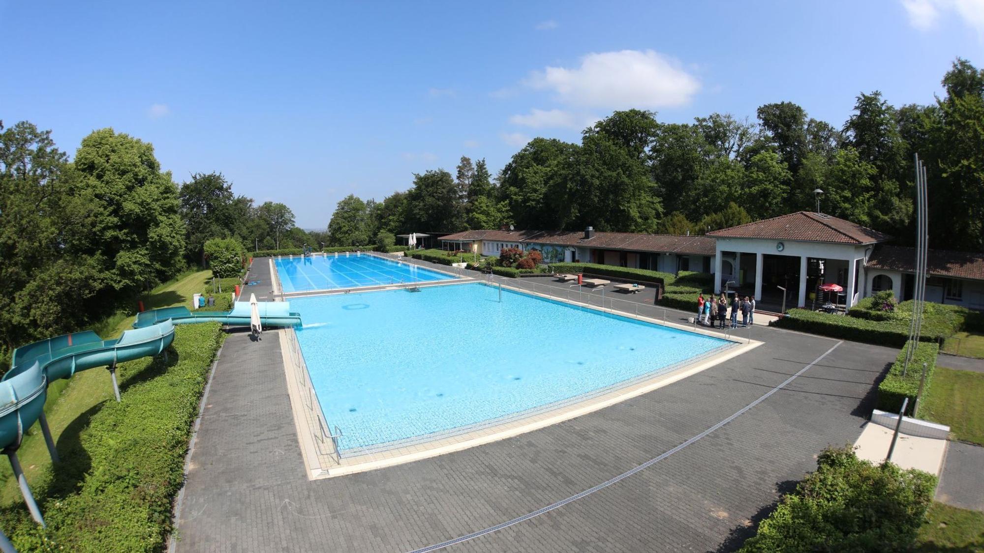 Mit Wasser gefüllt sind das Nichtschwimmer- und das 50-Meter-Schwimmerbecken. Links am Rand steht eine 65-Meter-Wasserrutsche, rechts ist das Eingangsgebäude zu sehen.