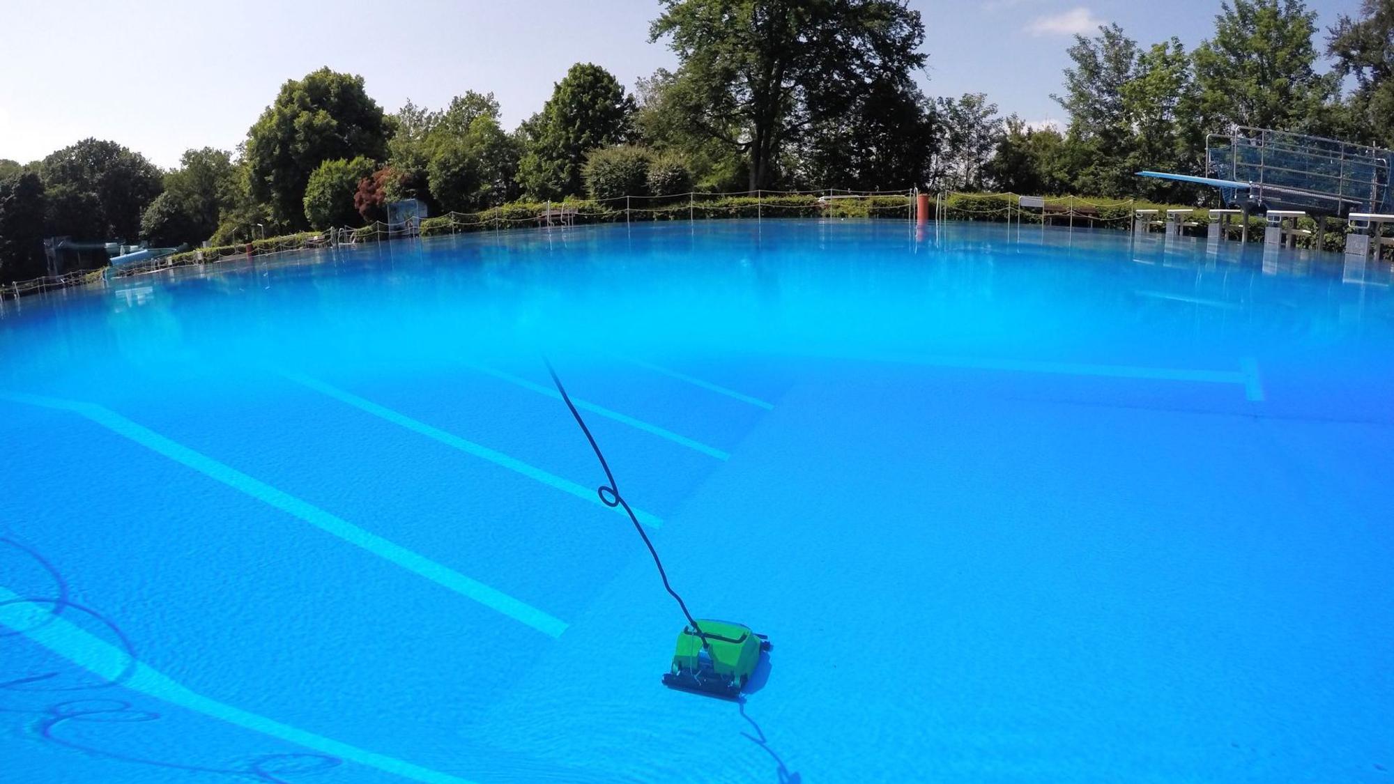 Im blauen Wasser zieht auf dem Boden des 50-Meter-Beckens eine gelbe Reinigungsmaschine ihre Bahnen. Am oberen Bildrand sind Bäume und Sträucher zu sehen, rechts am Rand ein Sprungbrett.