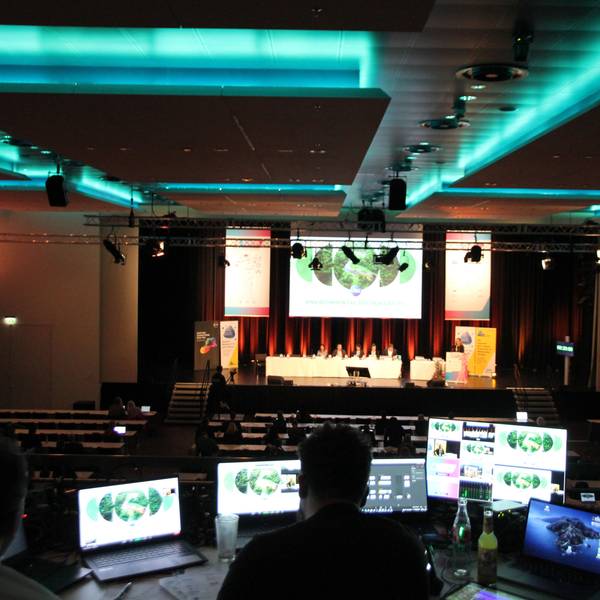 Zwei Männer an Computermonitoren auf der Empore eines großen Veranstaltungssaals mit einer Podiumsdiskussion auf der Bühne.