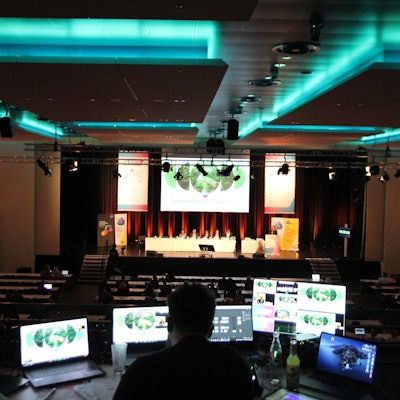 Zwei Männer an Computermonitoren auf der Empore eines großen Veranstaltungssaals mit einer Podiumsdiskussion auf der Bühne.