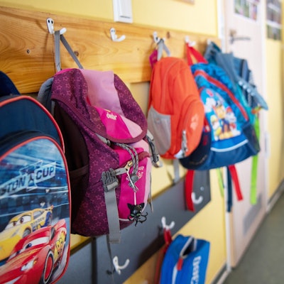 Das Foto zeigt die Garderobe in einer Kita. An den Haken hängen bunte Rucksäcke.